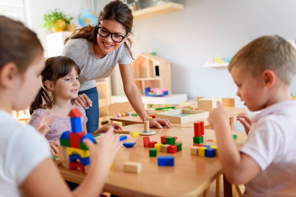 Kinder spielen am Tisch mit Bauklötzen. Eine Frau mit Brille steht und lächelt den Kindern zu.