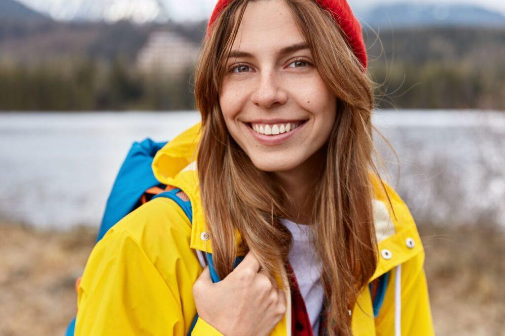 Lächelnde Frau in gelber wetterfester Outdoor-Jacke, roter Mütze und blauem Rucksack vor einer natürlichen Kulisse mit See und Bergen, ideal für Freizeitaktivitäten. Alpinestars-Inspiration für funktionelle und modische Outdoor-Bekleidung.