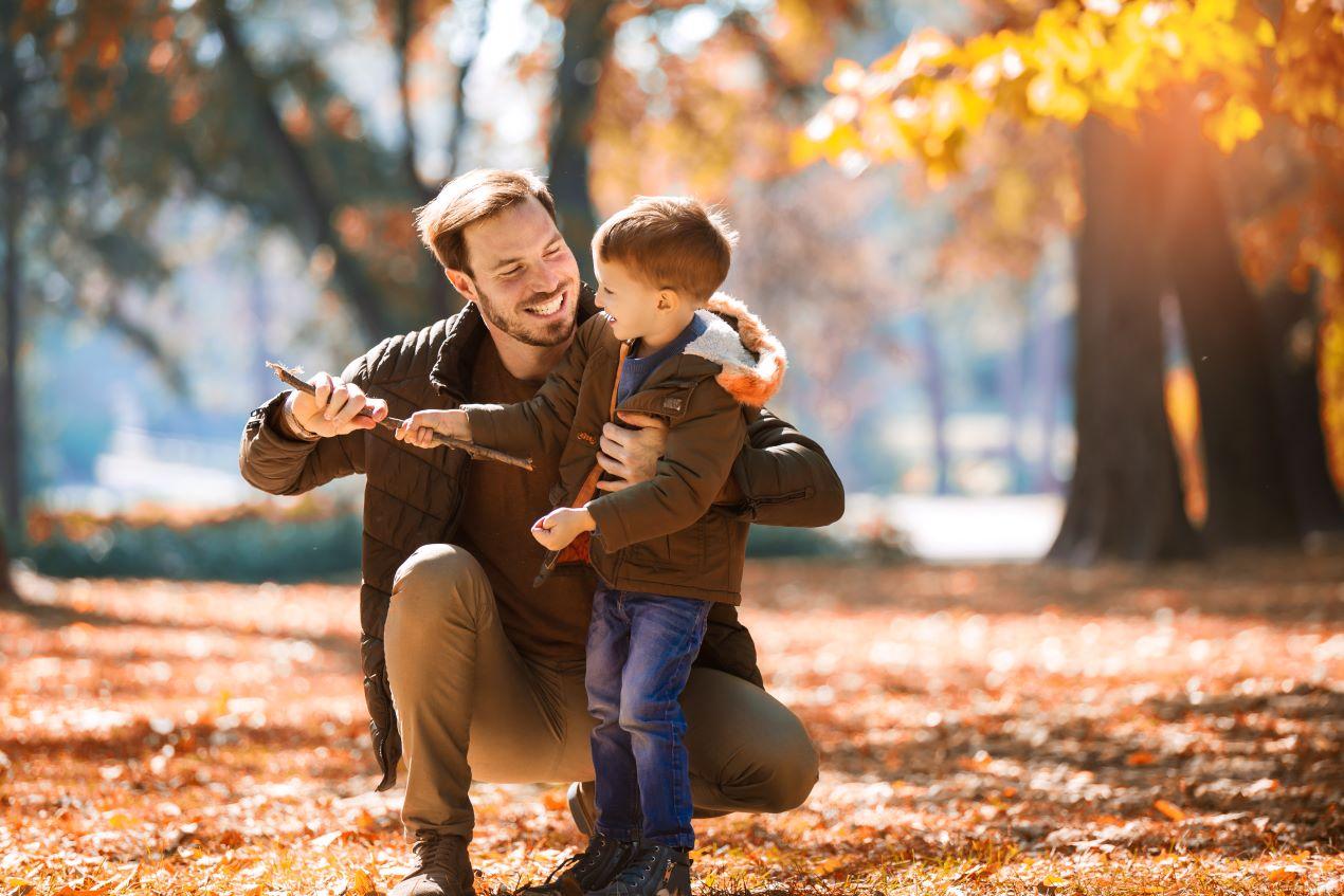 Vater und Sohn in brauner wetterfester Outdoor-Bekleidung spielen im Herbstwald mit buntem Laub. Beispiel für funktionale Kleidung für Eltern und Kinder. Alpinestars zeigt ähnliche Qualität für Motorradfahrer.