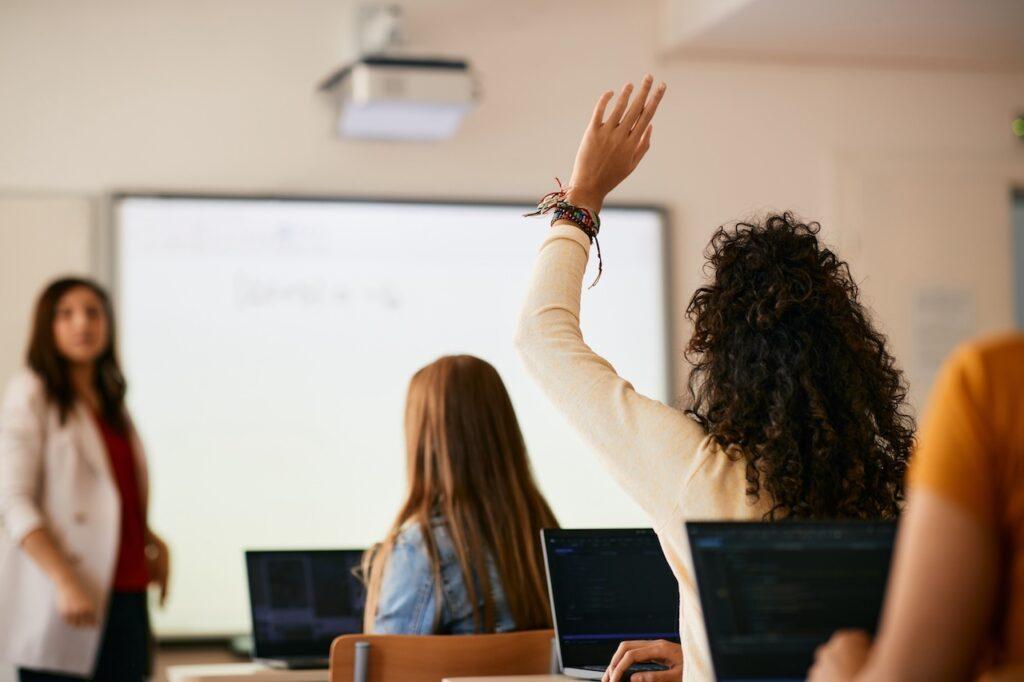 Eine Schülerin hebt im Unterricht die Hand, um eine Frage zu stellen, während die Lehrerin vor einer interaktiven Tafel unterrichtet.